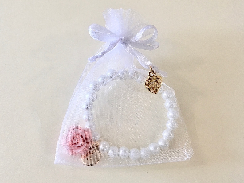 May include: A white pearl bracelet with a pink flower charm and a gold heart charm. The bracelet is packaged in a white organza bag with a white satin ribbon.