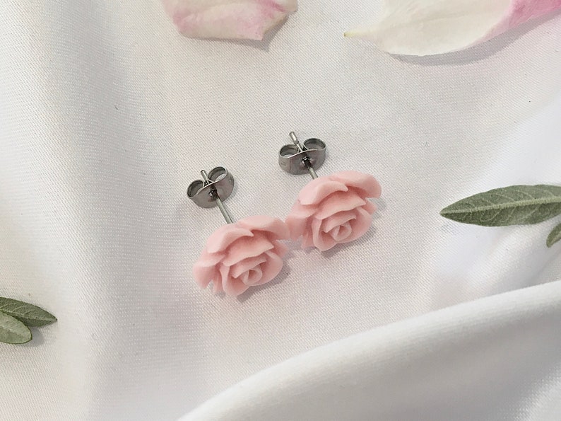 Op de afbeelding: Een paar zilveren oorbelletjes met roze rozenbloem accenten. De oorbelletjes liggen op een witte achtergrond.