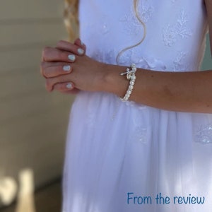 May include: A white pearl bracelet with a silver cross charm. The bracelet is worn on a person's wrist.