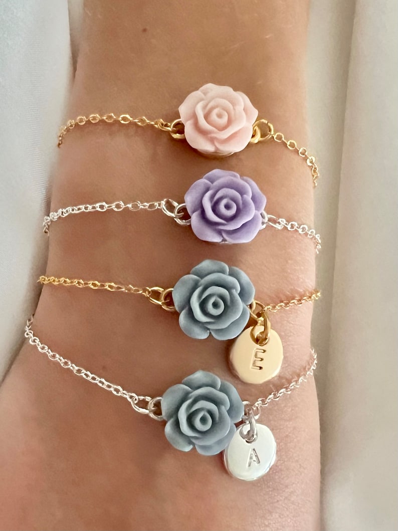 May include: Four delicate bracelets with silver and gold chains. Each bracelet features a different colored flower charm, including pink, purple, gray, and a silver charm with the letter 'E' and a gray charm with the letter 'A'.