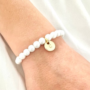 Puede incluir: Un brazalete de perlas blancas con un dije de oro con la letra 'C'.