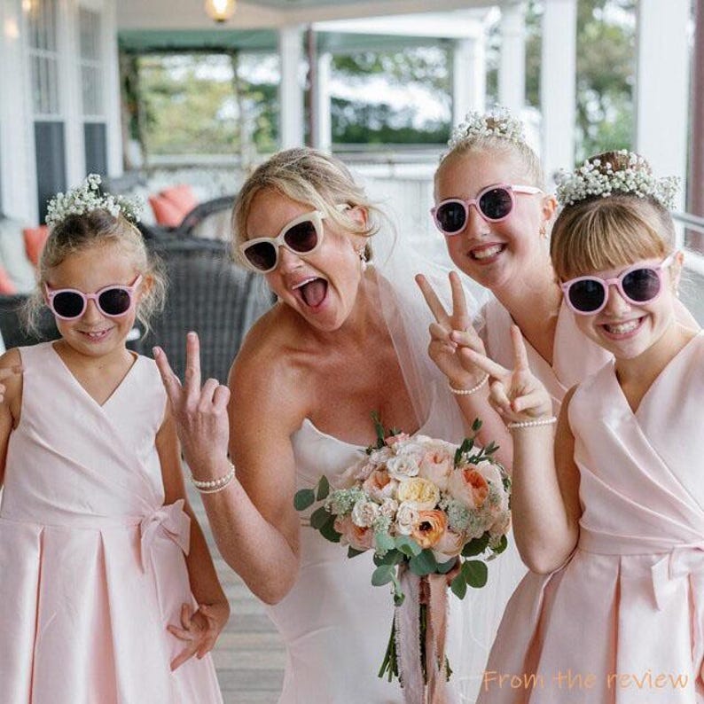 Puede incluir: Una novia con un vestido blanco posa con tres damas de honor que llevan vestidos rosas y gafas de sol. Las ni&ntilde;as hacen el signo de la paz y sonr&iacute;en. La novia lleva un ramo de flores.