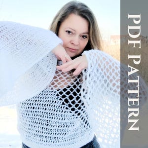 May include: A white crochet top with a lacy, open-weave design. The top has long, wide sleeves and a loose, flowing fit. The model is wearing the top over a black tank top.
