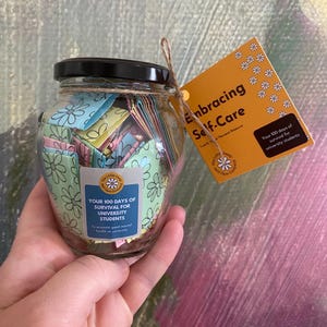 May include: A glass jar with a black lid, filled with colorful paper notes, tied with twine. A yellow tag reads "Embracing Self-Care" and "Your 100 days of survival for university students". The jar is decorated with floral patterns.