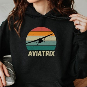 May include: Black hoodie with a vintage sunset graphic and a silhouette of a small airplane. The text "AVIATRIX" is printed below the graphic.