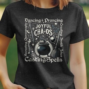 Puede incluir: Una camiseta negra con un diseño gráfico blanco que presenta un caldero, estrellas y el texto "Dancing & Prancing", "Joyful Chaos", "Creating Inside Jokes" y "Casting Spells".