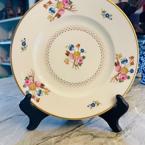 May include: A round white porcelain plate featuring a gold rim and a central floral bouquet of pink, yellow, and blue flowers. The plate is decorated with a gold lattice border pattern and additional floral sprays around the edge of the rim.