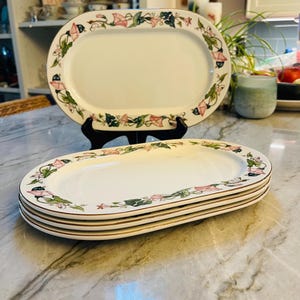 May include: A set of four white oval ceramic serving platters featuring a decorative border of pink morning glory flowers and green leaves with a thin gold rim. One platter is displayed upright on a black stand, while three are stacked below.