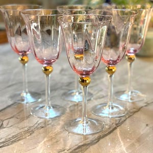 May include: A set of six clear glass goblets featuring a subtle pink tint at the base of the bowl. Each stem includes a prominent gold-colored spherical accent located just below the bowl, supported by a clear circular glass base.