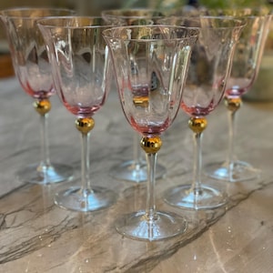 May include: A set of six clear glass wine glasses with a subtle pink tint at the base of the bowl. Each glass features a distinct gold-colored spherical accent on the stem, connecting the bowl to the clear glass base, with etched floral patterns.