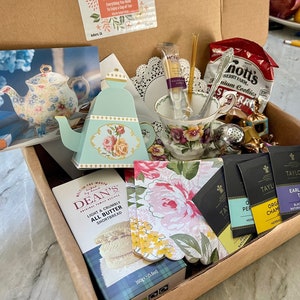 May include: A brown cardboard box filled with a tea kit. The kit includes a teapot, teacups, tea bags, cookies, and shortbread. The box also includes a card that says "The Tea Kit" and "Everything You Need To Enjoy a Cup of Tea".