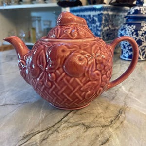 May include: A dusty rose ceramic teapot featuring a raised relief pattern of woven basket textures, leaves, and various fruits. The teapot includes a matching lid with a small fruit-shaped finial, a curved handle, and a short, tapered spout.