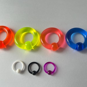 Puede incluir: Colección de coloridas joyas para el cuerpo. Cuatro grandes anillos cautivos de acrílico en naranja, verde neón, rosa y azul. Debajo, tres anillos más pequeños en blanco, negro y morado, sobre fondo claro.