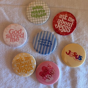 May include: Seven circular pinback buttons featuring various designs and text. One is green gingham with 'MAKE HEAVEN CROWDED', one red with white dots and 'ask me about Jesus', one pink with floral patterns and 'God saved me', one blue striped with 'what if my purpose is to LOVE GOD?', one yellow with 'JESUS saved my life', one orange with white dots and 'ask me about Jesus', and one pink with 'JESUS LOVES YOU'.