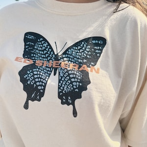 Peut inclure: Sweat-shirt crème avec un grand motif de papillon. Le papillon est noir et bleu, avec le nom "ED SHEERAN" en texte couleur cuivre. Les ailes du papillon sont remplies des mots "ED SHEERAN".