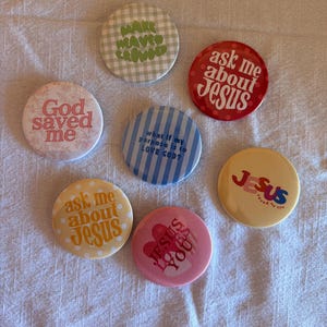 May include: A collection of seven round pinback buttons with various designs and text. The buttons feature phrases like "God saved me," "Ask me about Jesus," and "Jesus loves you." Colors include red, pink, yellow, and blue.
