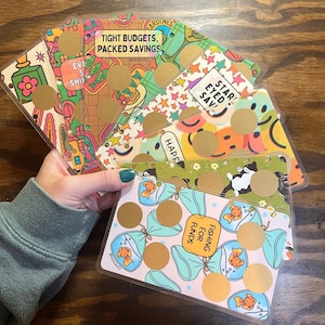 May include: A hand holds a collection of colorful, patterned budget cards. Each card features unique designs, including smiley faces, fish, and text like "Tight Budgets, Packed Savings." Gold scratch-off circles are present on each card.