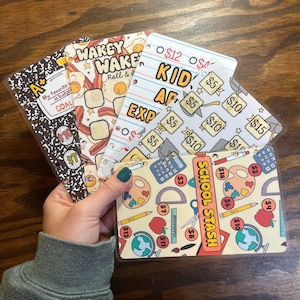 May include: A hand holds several budget binder inserts. Designs include a composition notebook, bacon and eggs, lined paper, and school supplies. Text includes "GOAL", "WAKEY WAKE", "KID EXP", and "SCHOOL STASH", with dollar amounts.