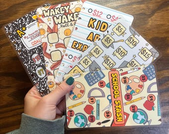 May include: A hand holds several budget binder inserts. Designs include a composition notebook, bacon and eggs, lined paper, and school supplies. Text includes "GOAL", "WAKEY WAKE", "KID EXP", and "SCHOOL STASH", with pound amounts.