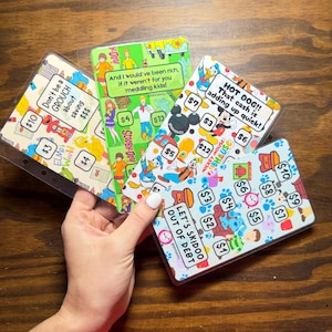 May include: A collection of colorful scratch-off savings cards with various designs and text. The cards feature characters like Elmo, Scooby-Doo, and Mickey Mouse, with phrases like "Don't be a GROUCH" and "LET'S SKIDOO OUT OF DEBT".