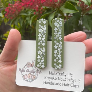 May include: Two green hair clips with white floral designs. The clips are on a white card with the text "NelsCraftyLife Etsy/IG: NelsCraftyLife Handmade Hair Clips".