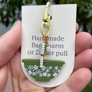 May include: Handmade green and white floral bowl charm or zipper pull with white beads and gold accents. The charm is attached to a gold metal clasp.