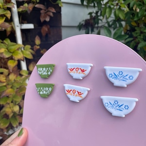 May include: A collection of miniature ceramic bowls with floral designs. The bowls are in various colors, including white, green, and pink, and are arranged on a pink surface. The bowls feature detailed patterns and are likely decorative items.