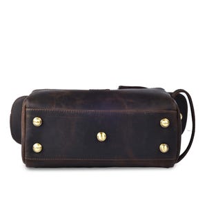 May include: A dark brown leather duffel bag with gold-colored metal studs on the bottom. The bag has a rectangular shape with rounded corners and a handle on the side. The leather has a distressed appearance.