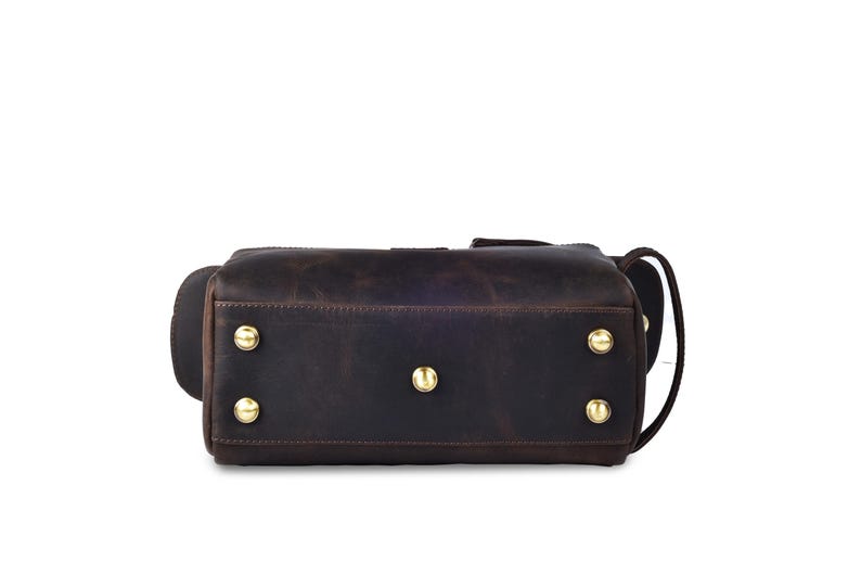 May include: A dark brown leather duffel bag with gold-colored metal studs on the bottom. The bag has a rectangular shape with rounded corners and a handle on the side. The leather has a distressed appearance.