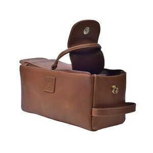 May include: A brown leather toiletry bag with a handle and a zippered main compartment. The bag has a flap closure with a magnetic snap and a small handle on the side. The bag has a small embossed logo on the front.