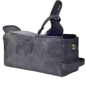 May include: A dark gray leather toiletry bag with a zipper closure and a handle. The bag has a rectangular shape with a flap on top and a small handle on the side. The bag has the text "LEATHER & LANCE" on the front.