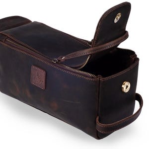 May include: A dark brown leather toiletry bag with a rectangular shape and a zippered main compartment. The bag has a flap closure, a handle, and a small logo patch. The bag is open, revealing the interior.