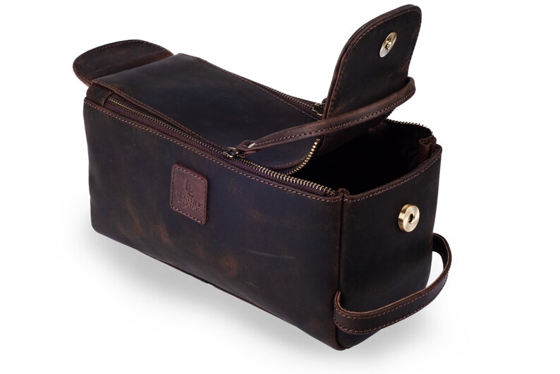 May include: A dark brown leather toiletry bag with a rectangular shape and a zippered main compartment. The bag has a flap closure, a handle, and a small logo patch. The bag is open, revealing the interior.