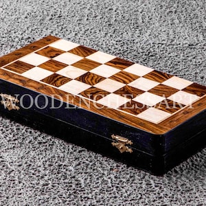 Könnte beinhalten: Ein Schachbrett aus Holz mit einem schwarzen und braunen Holzeinlege-Design. Das Brett ist geschlossen und hat eine schwarze Außenseite mit goldenen Scharnieren. Das Brett ist mit "WOODEN CHESS ART" beschriftet.