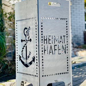 Könnte beinhalten: Ein silberner Metall-Feuerkorb mit Anker-Design und den ausgeschnittenen Worten "HEIMAT HAFEN". Der Feuerkorb hat eine rechteckige Form mit einem Griff oben und ist für den Außenbereich konzipiert.