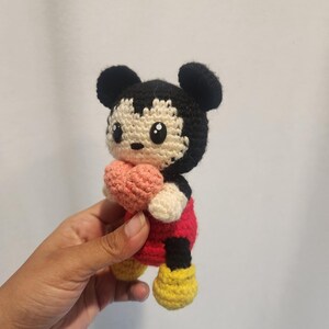 Op de afbeelding: Gehaakte Mickey Mouse pop met een roze hart. De pop heeft zwarte oren, een zwart en cr&egrave;me gezicht, een rode body, cr&egrave;me handen en gele schoenen. Een handgemaakt, grillig speelgoed.