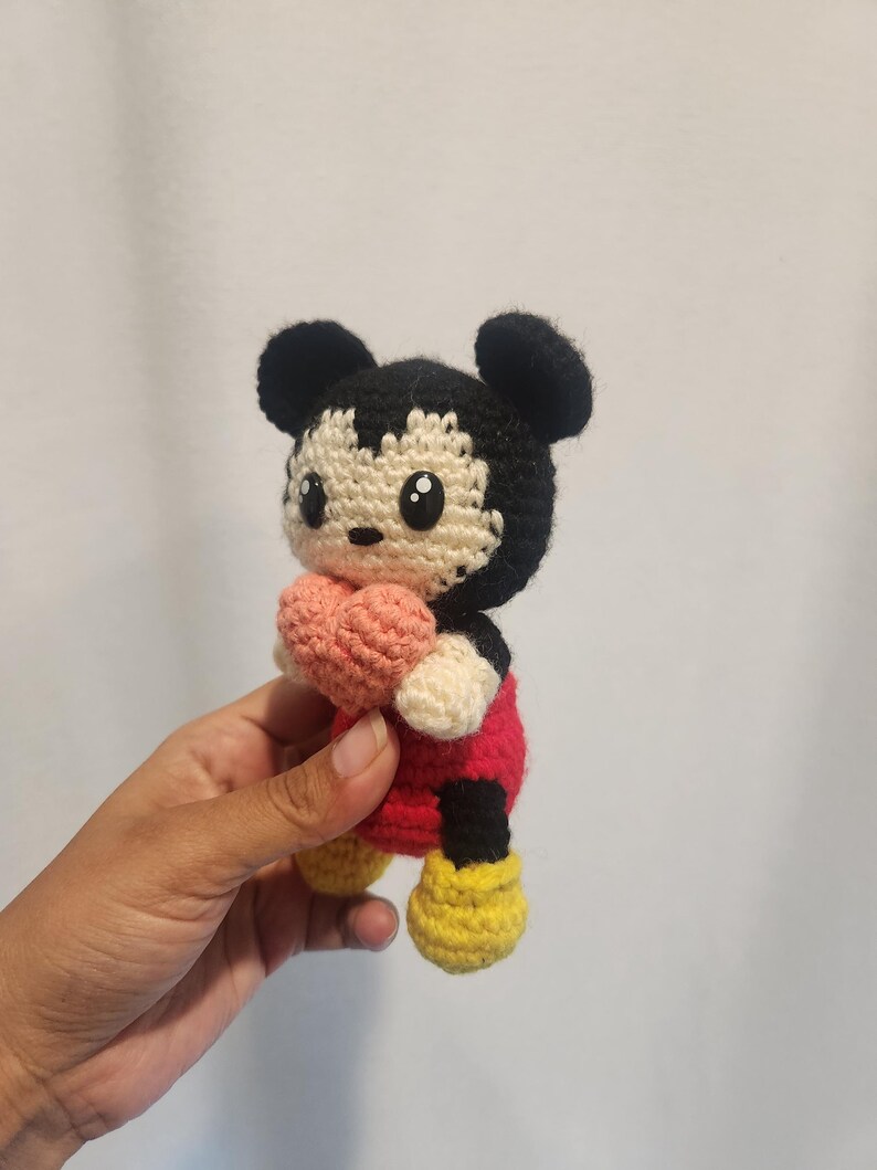 Op de afbeelding: Gehaakte Mickey Mouse pop met een roze hart. De pop heeft zwarte oren, een zwart en cr&egrave;me gezicht, een rode body, cr&egrave;me handen en gele schoenen. Een handgemaakt, grillig speelgoed.