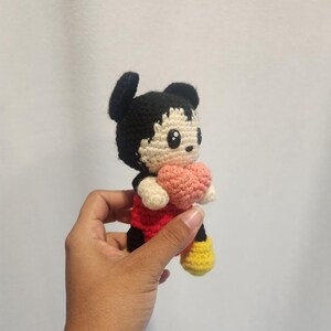 Op de afbeelding: Handgemaakte gehaakte Mickey Mouse-pop, met een zwart hoofd en oren, een rood shirt, een zwarte broek, gele schoenen en een roze hart. De pop is gemaakt van garen en heeft een schattig, cartoonachtig ontwerp.