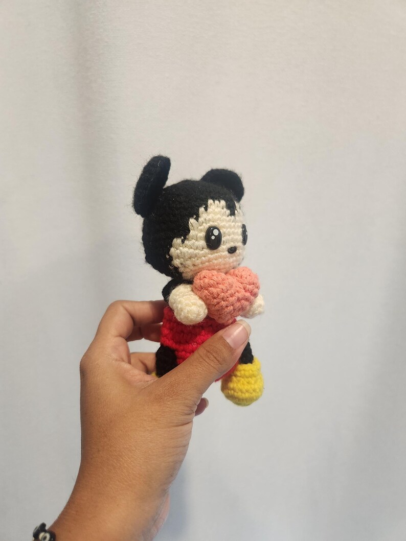 Op de afbeelding: Handgemaakte gehaakte Mickey Mouse-pop, met een zwart hoofd en oren, een rood shirt, een zwarte broek, gele schoenen en een roze hart. De pop is gemaakt van garen en heeft een schattig, cartoonachtig ontwerp.