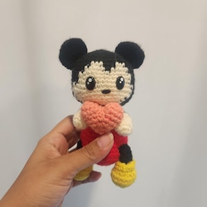 Op de afbeelding: Gehaakte Mickey Mouse pop met een roze hart. De pop heeft zwarte oren, een zwart en cr&egrave;me gezicht, een rode body, zwarte shorts en gele schoenen. Het handgemaakte speelgoed is een schattig en knuffelbaar item.