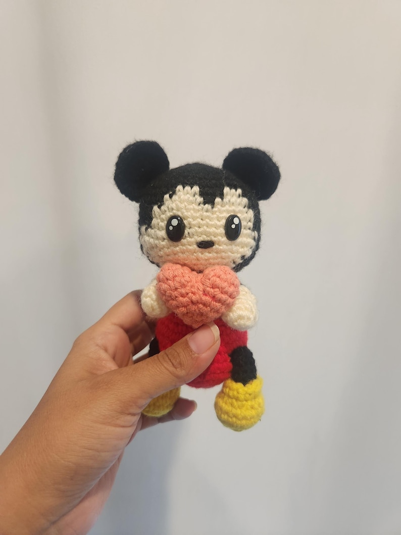 Op de afbeelding: Gehaakte Mickey Mouse pop met een roze hart. De pop heeft zwarte oren, een zwart en cr&egrave;me gezicht, een rode body, zwarte shorts en gele schoenen. Het handgemaakte speelgoed is een schattig en knuffelbaar item.