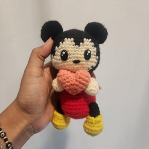 Op de afbeelding: Gehaakte Mickey Mouse pop met een roze hart. De pop heeft zwarte oren, een cr&egrave;mekleurig gezicht, een rode body, zwarte shorts en gele schoenen. De pop heeft grote, zwarte ogen en is handgemaakt.