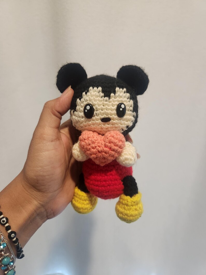Op de afbeelding: Gehaakte Mickey Mouse pop met een roze hart. De pop heeft zwarte oren, een cr&egrave;mekleurig gezicht, een rode body, zwarte shorts en gele schoenen. De pop heeft grote, zwarte ogen en is handgemaakt.