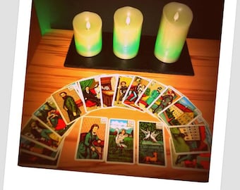 May include: A tarot card reading spread with 14 cards arranged in a semi-circle. The cards are colorful and feature illustrations of people and scenes. Three green candles are lit and placed on a black slate surface in front of the cards.
