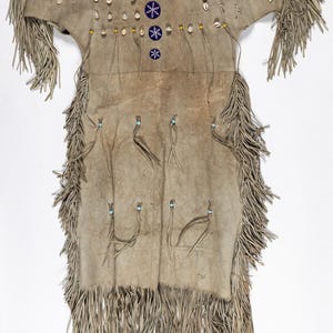 Op de afbeelding: Een bruin leren kledingstuk met franjes, versierd met schelpparels en blauwe en rode geometrische ontwerpen.