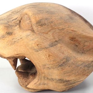 May include: A wooden sculpture of a big cat's head, with its mouth open, showing a single tooth. The sculpture is made of light brown wood with darker streaks. The details of the cat's face are visible.