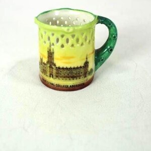 Op de afbeelding: Een kleine keramische mok met een groene handgreep en rand. De mok heeft een geel en bruin beeld van een gebouw en de tekst "MARISCHAL COLLEGE ABERDEEN". De mok heeft een traliewerkontwerp rond de body.