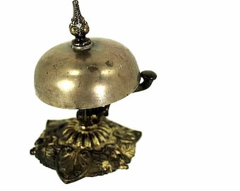 May include: A small, antique brass bell with a domed top and ornate base. The bell has a small handle and a decorative finial. The base is shaped like a flower with detailed leaf patterns. The bell is likely made of brass or a similar metal.