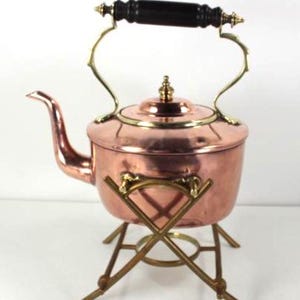 May include: A copper tea kettle with a black wooden handle and brass detailing. The kettle is supported by a brass stand. The spout curves elegantly to the left. A classic kitchenware item.