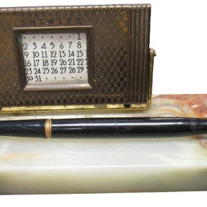 May include: A vintage desk set made of marble with a gold-toned calendar and a black fountain pen. The calendar has a small window that displays the date and the days of the week. The pen has a black barrel and a gold-toned nib.